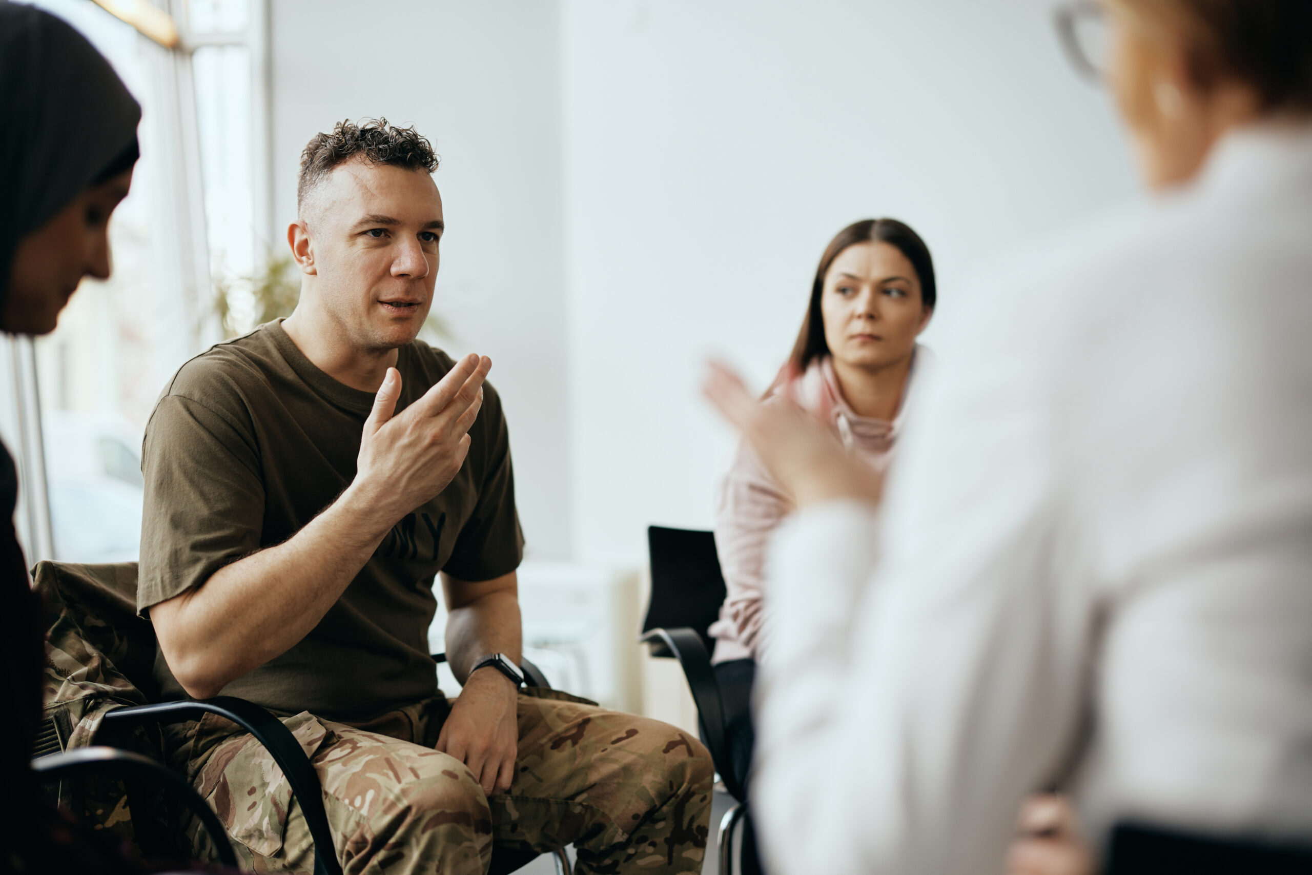 A man in military camouflage pants, likely a Marine, engages in a group discussion. He gestures with his hand while speaking. Two women sit attentively with him in the bright room, their expressions indicating their keen interest in addressing mental health treatment needs.