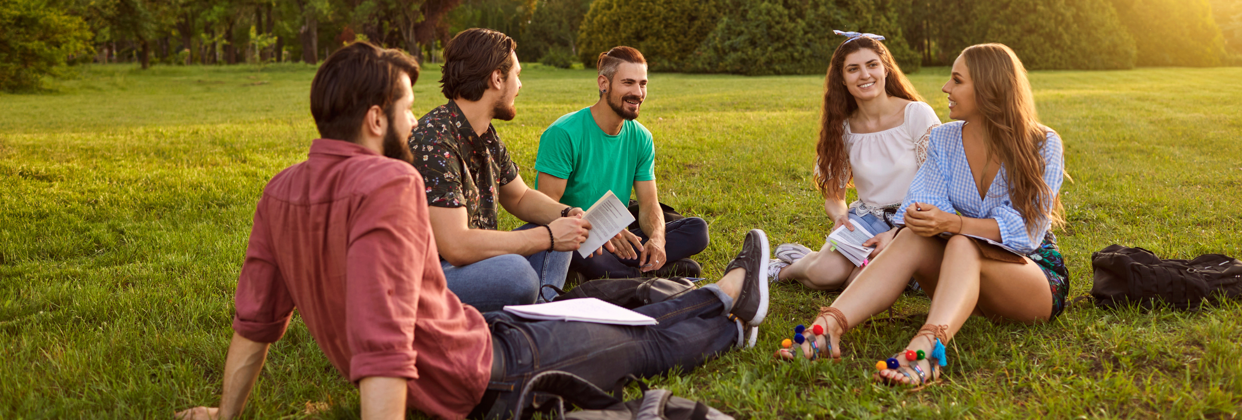 A group of five people sits on the grass in a park, engaged in a lively conversation about an Intensive Outpatient Program for Mental Health. Relaxed and holding books, they enjoy the sunny day amid trees and bushes, clearly appreciating both nature and their supportive dialogue.