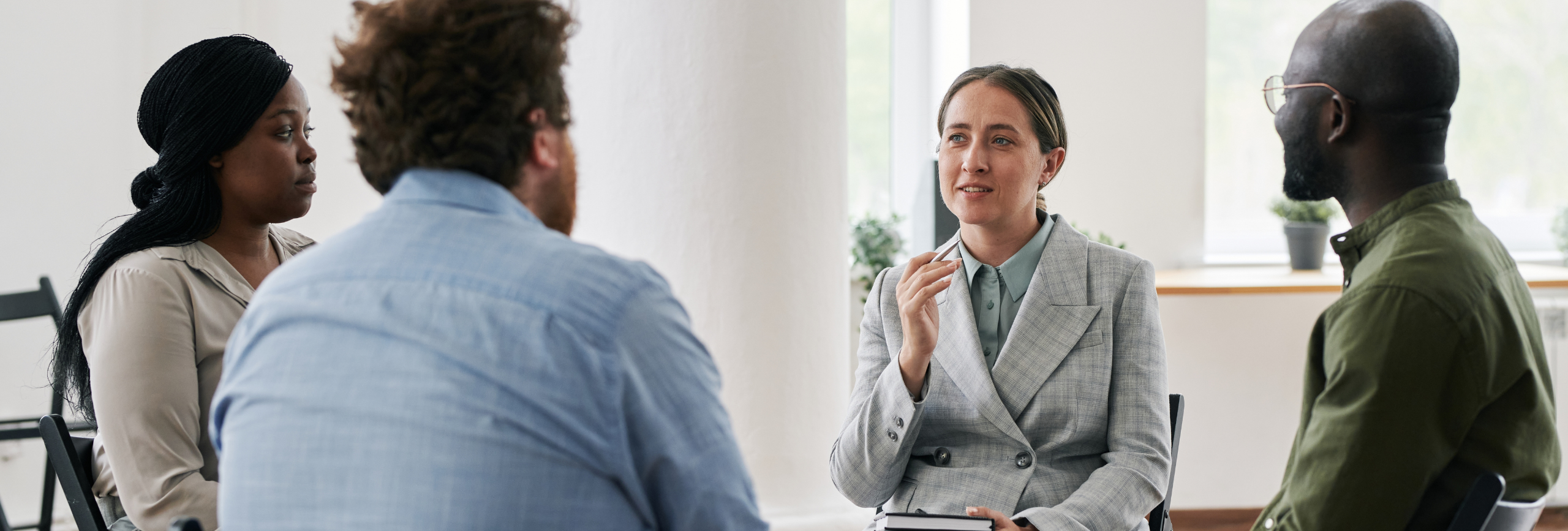 A group of four people sits in a circle having a discussion on depression treatment in Chicago. One person in a gray suit speaks while gesturing with a hand as the others casually listen. Theyre in a bright room with large windows and potted plants in the background.