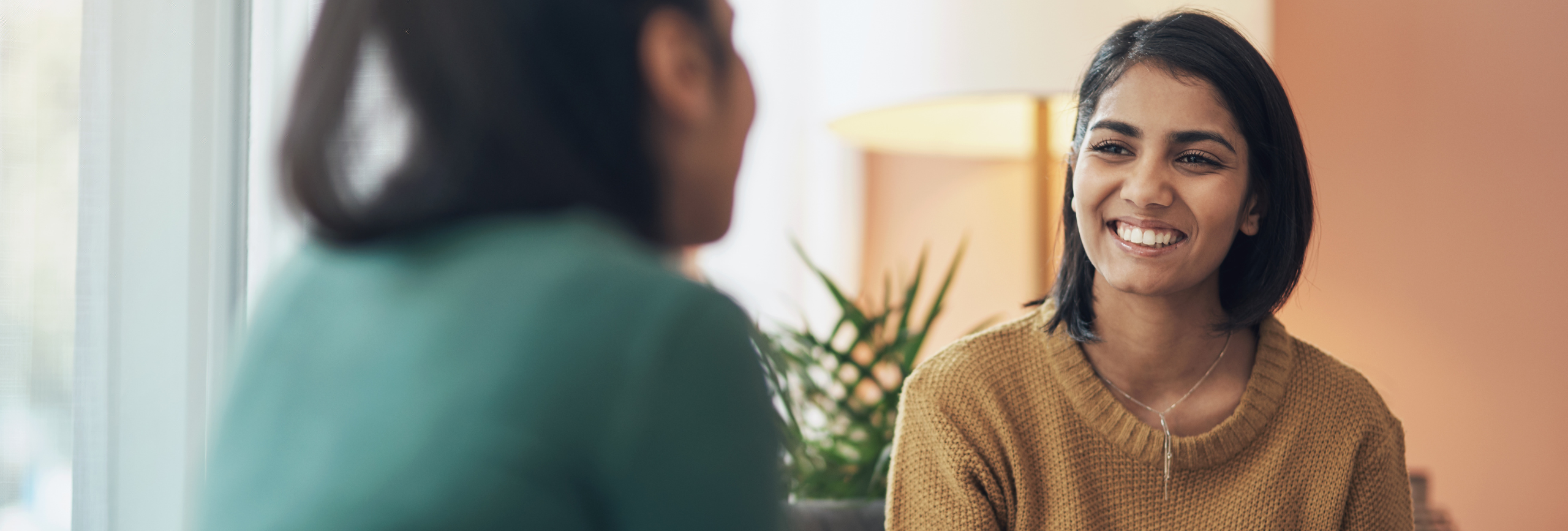 Two women sit indoors facing each other and smiling warmly, reflecting a supportive atmosphere often found in womens mental health treatment. One, in focus, wears a mustard sweater and necklace; a plant and lamp are visible in the background.