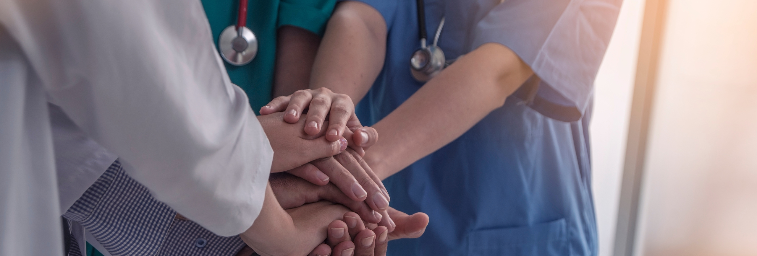 A group of healthcare workers wearing scrubs and stethoscopes stack their hands in a show of teamwork and unity, highlighting their commitment to supporting each other and advancing mental health treatment.