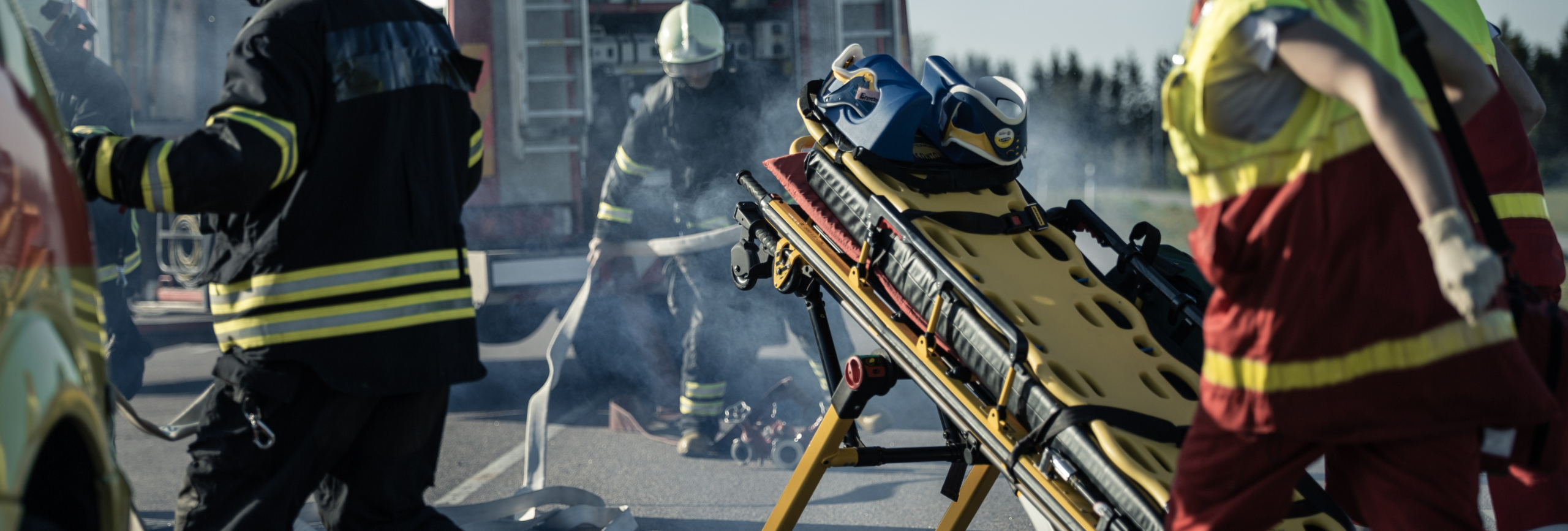 Firefighters and paramedics respond to an emergency scene, with a stretcher in the foreground and smoke rising around them, highlighting the intense situations that make First Responders mental health treatment so important.
