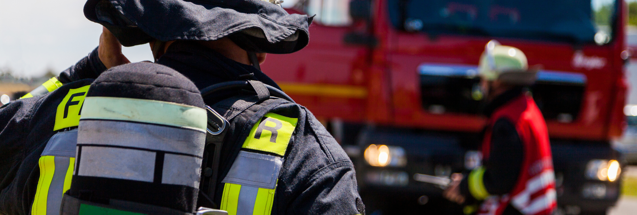 A firefighter in full gear stands in the foreground, seen from behind, while another—focused on firefighters mental health treatment—wears a red vest near a fire truck in the background.