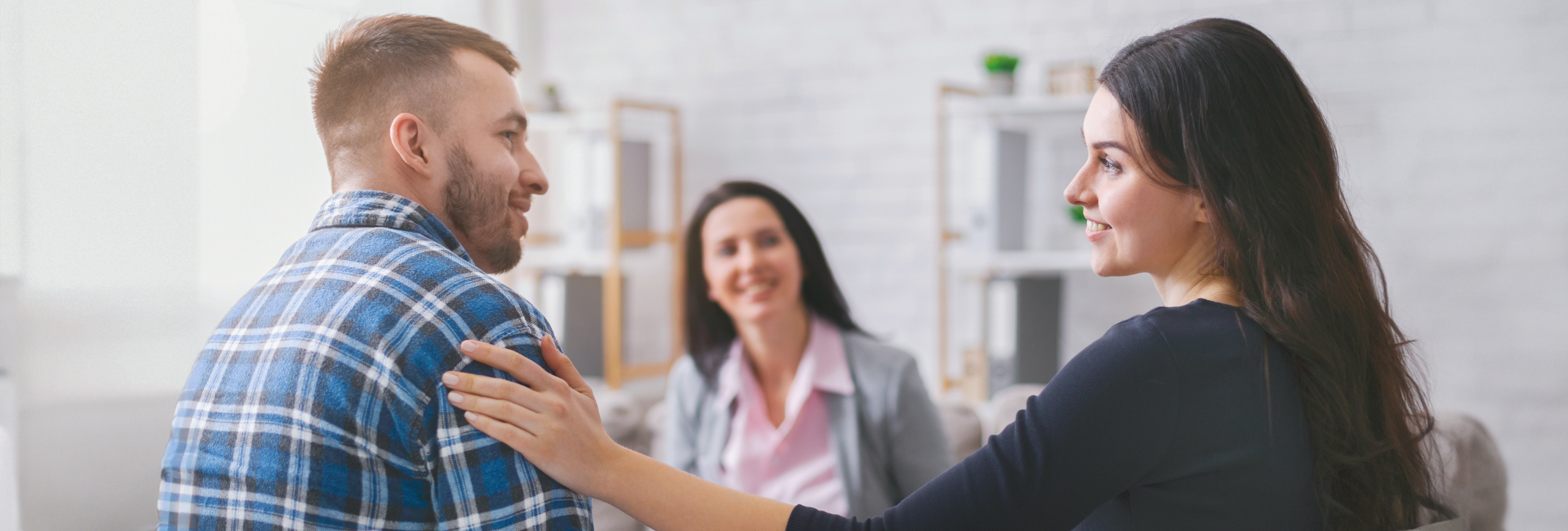 A man and woman sit facing each other, smiling as the woman touches the mans shoulder. In a bright room, a third woman—possibly offering Family Therapy in Chicago—sits in the background, also smiling warmly.