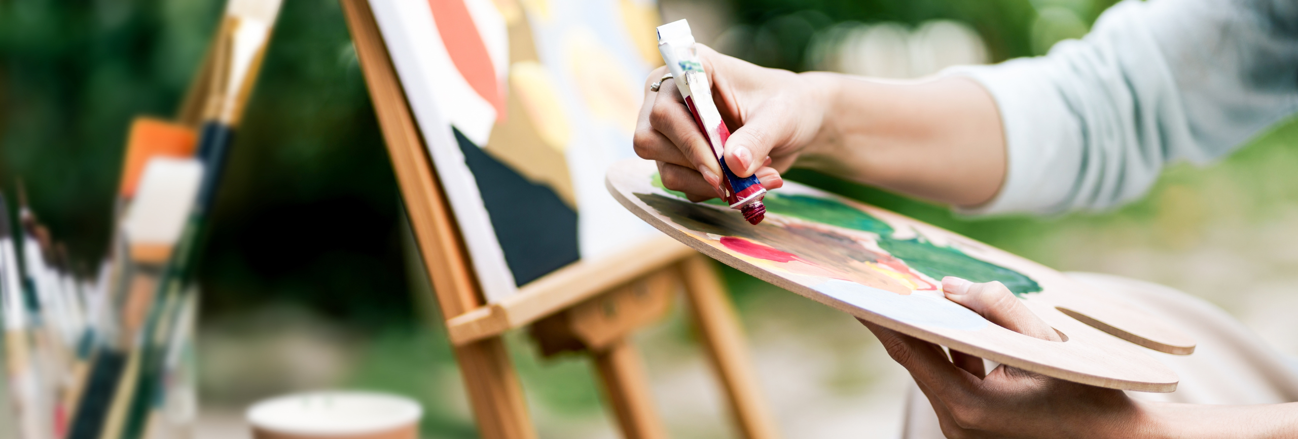 A person mixes paint on a wooden palette with a brush, while an easel holding a colorful canvas stands in the background outdoors—capturing the calming spirit of Art Therapy in Chicago.