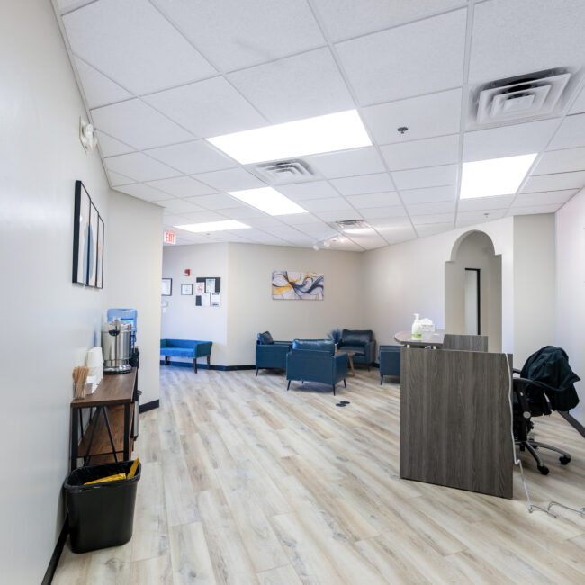 A bright, modern office waiting area welcomes visitors for a tour, featuring light wood flooring, white walls, a reception desk on the right, water station on the left, blue chairs and couches in back, and artwork on the walls.