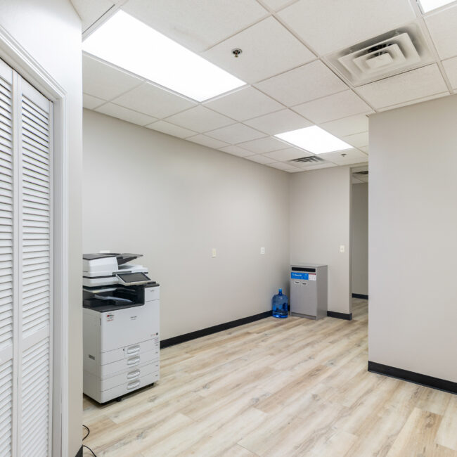 Take a virtual tour of this clean, modern office space featuring light wood flooring, off-white walls, fluorescent lights, a printer, water dispenser with blue jugs in the corner, and white louvered closet doors to the left.