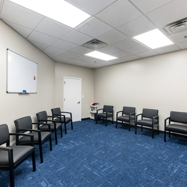 A brightly lit room with blue carpet, beige walls, and black chairs arranged in a circle. There is a whiteboard on the left wall, a small table with magazines—a welcoming space for your tour—plus a door at the back.