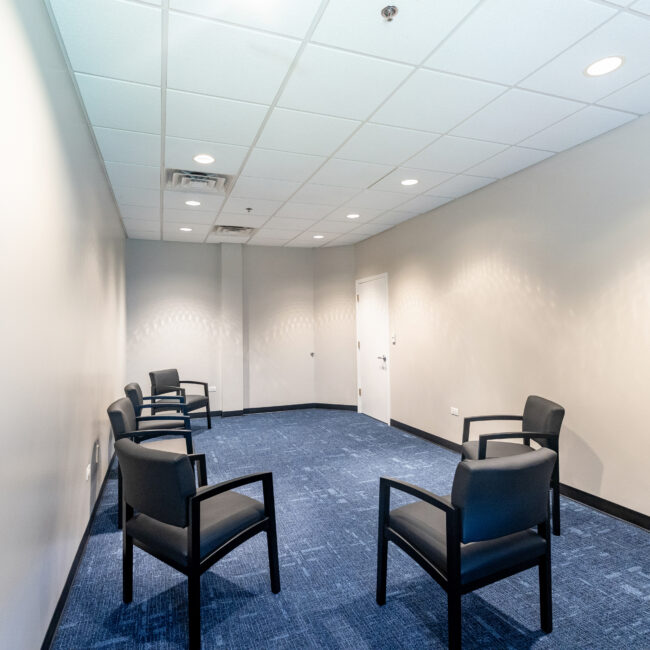 A small, plain room with white walls, a blue carpet, a white ceiling with recessed lights, and six black armchairs arranged in a circle—ideal for a group tour or meeting. A closed white door is visible at the far end of the room.