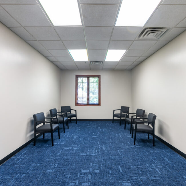 A small tour-ready room with blue carpet, white walls, six black chairs spaced along the side walls, a window with blinds at the far end, and fluorescent ceiling lights. The room appears empty and tidy.