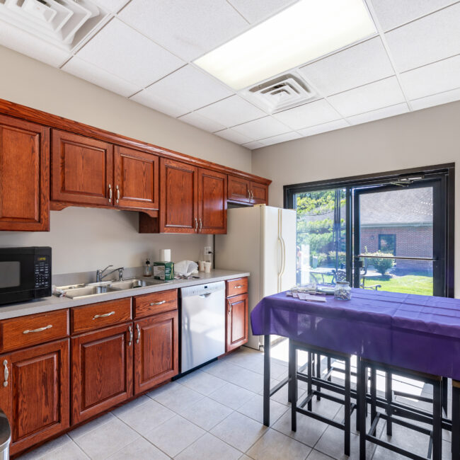 Take a tour of this inviting break room featuring wooden cabinets, sink, microwave, refrigerator, and dishwasher. A table with a purple cloth sits at the center, while windows reveal an outdoor patio and lush greenery beyond.