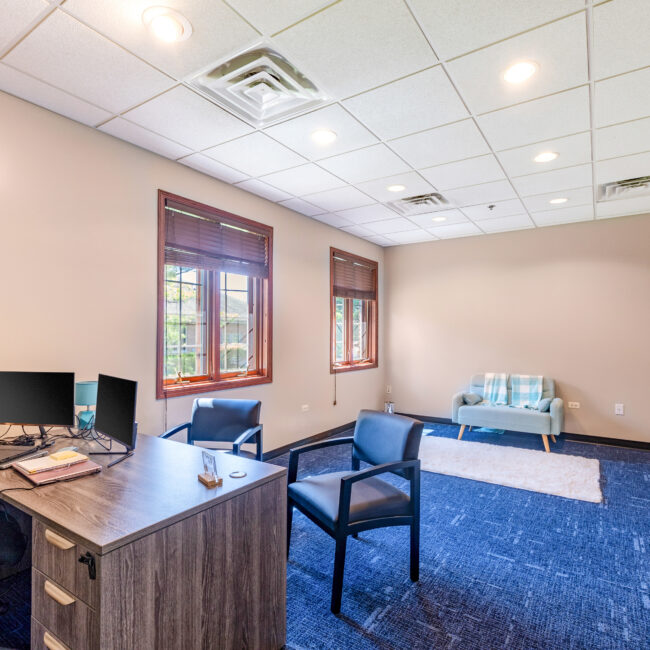 Take a tour of this bright office featuring a wooden desk, dual monitors, two chairs, and a cozy couch. Large windows fill the space with natural light, while dark blue carpet and light beige walls create a welcoming atmosphere.
