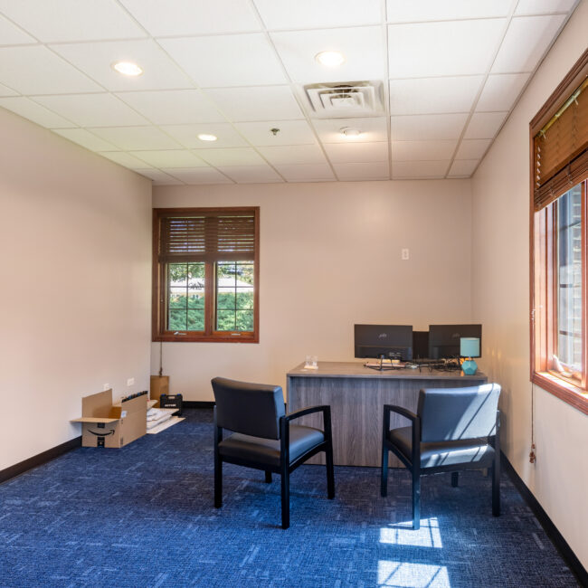 Take a quick tour of this small office with beige walls, blue carpet, a wooden desk with two monitors, two black chairs, and windows letting in natural light. Cardboard boxes and cables are on the floor near one wall.