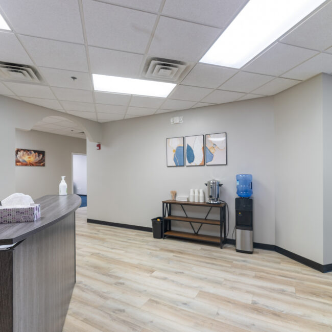 A clean, modern office waiting area with light wood flooring, a curved reception desk, wall art, a coffee station, and a water cooler awaits your tour under bright ceiling lights and light gray walls.