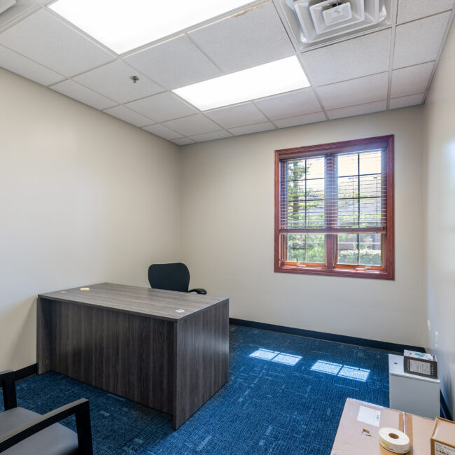 Take a quick tour of this small office featuring a wooden desk, black office chair, two guest chairs, blue carpet, cream walls, a window with wooden trim, and fluorescent lights on the ceiling.
