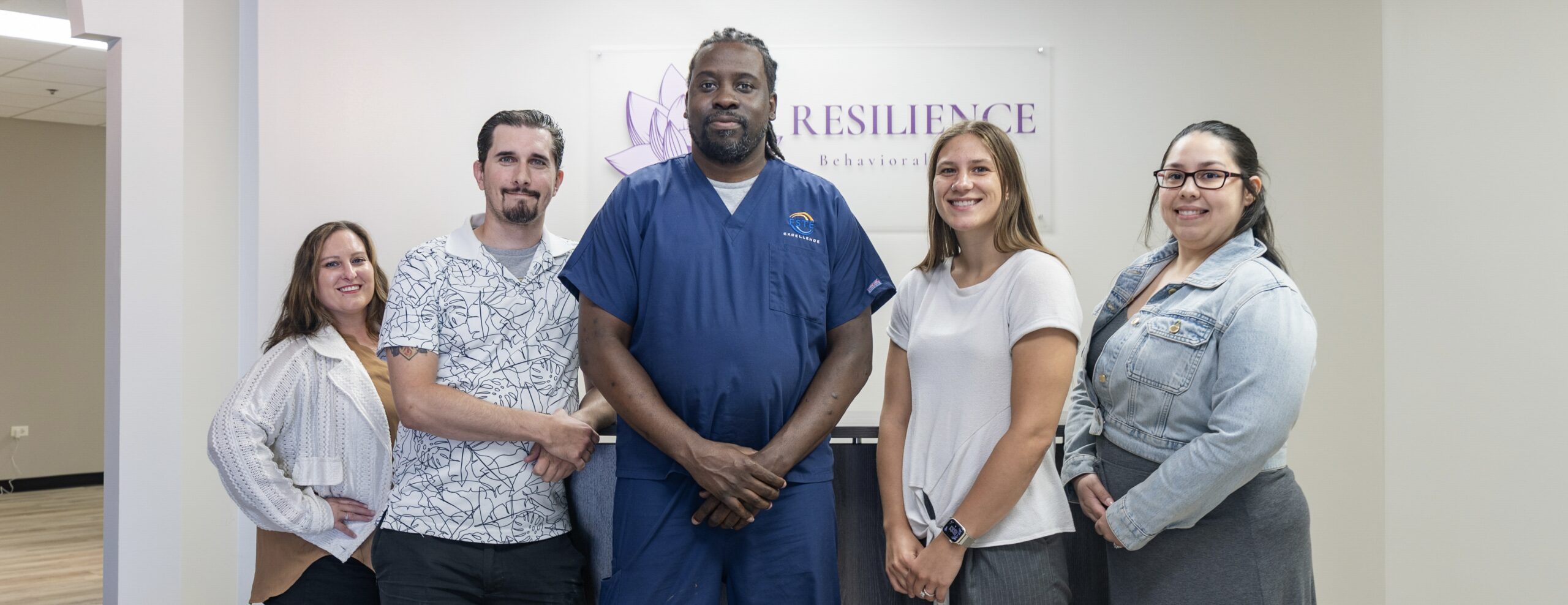 Meet the staff of Resilience BH: five people stand side by side, smiling at the camera in front of a sign that reads Resilience Behavioral. Four wear casual clothes while one is in medical scrubs.