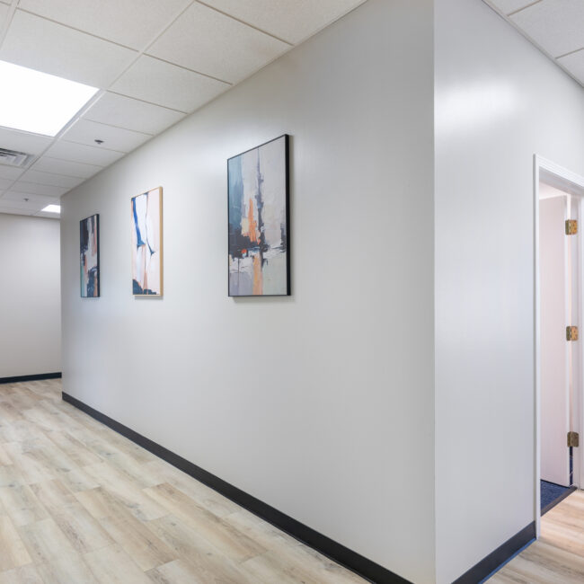 A clean, modern hallway with light wood flooring, white walls, and three abstract paintings invites you along a bright tour toward a door leading to another room. Ceiling lights provide vibrant illumination throughout the space.