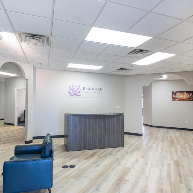 Modern office lobby with light wood floors, gray walls, a dark reception desk, and two blue chairs. The sign behind the desk reads RESILIENCE. Take a tour through the arched doorway to see artwork displayed in the background.