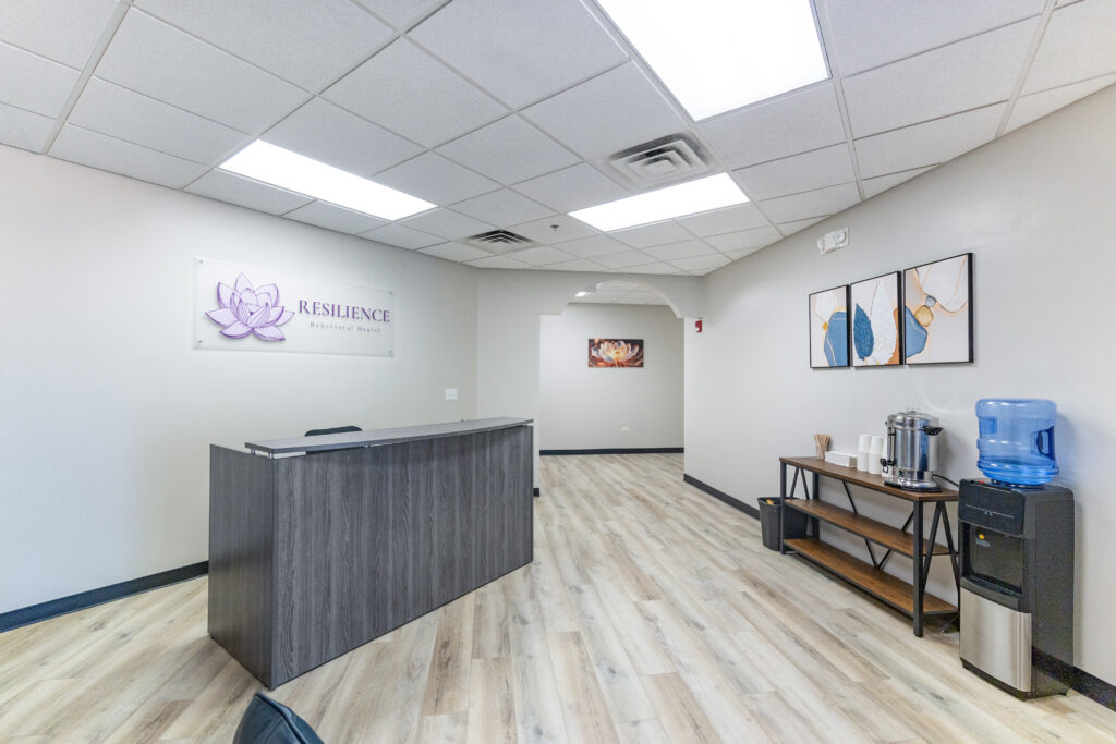 Tour our inviting reception area featuring a dark wooden desk, wall art, a Resilience Behavioral Health sign with a purple lotus, plus a refreshment table with coffee supplies and a water cooler on light wood flooring.