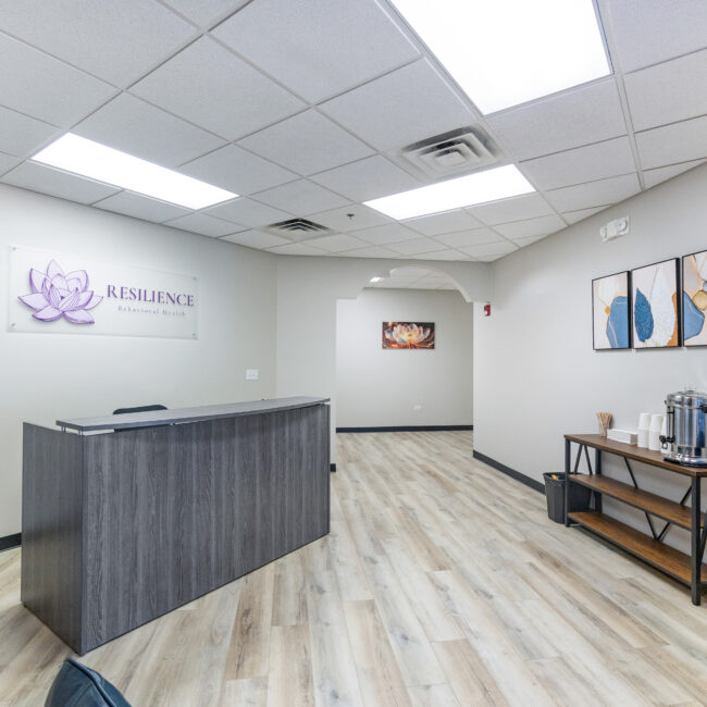 Tour our inviting reception area featuring a dark wooden desk, wall art, a Resilience Behavioral Health sign with a purple lotus, plus a refreshment table with coffee supplies and a water cooler on light wood flooring.