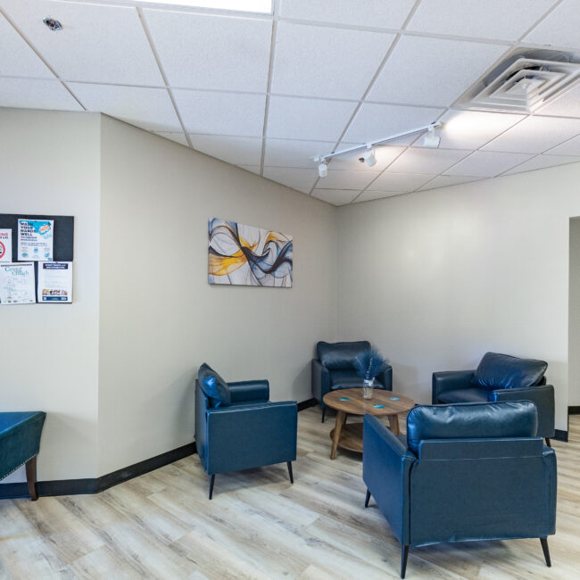 A waiting area with four blue armchairs around a small round table, a teal bench, wall art, and flyers on a beige wall. The light wood flooring and open doorway invite you to explore further—perfect for starting your tour of the space.