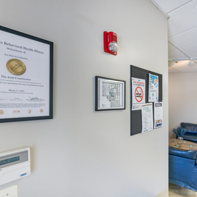 A framed accreditation certificate hangs on a wall next to safety signs and a thermostat, inviting visitors to tour the well-lit office space where blue chairs and small tables furnish the waiting area in the background.