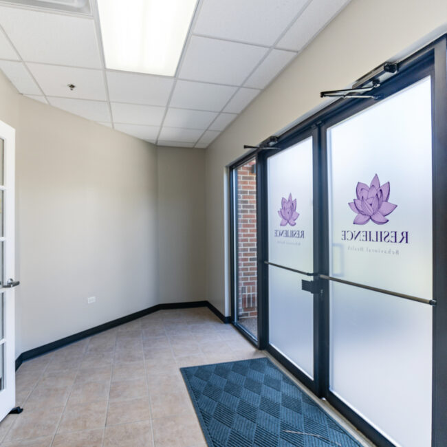 Take a tour of this bright office entrance with glass double doors featuring frosted panels and purple lotus logos, a glass-paneled door to the left, tiled flooring, and sunlight streaming in from outside.
