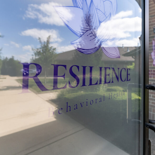 A glass door with Resilience Behavioral Health and a lotus flower logo, reflecting trees, a sidewalk, and a brick building under a blue sky with clouds—inviting you to tour the welcoming space within.