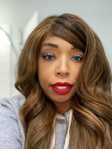 Meet The Staff at Resilience BH: A woman with long, wavy brown hair, bright blue eyes, and bold red lipstick, wearing a gray hoodie, takes a selfie in a well-lit indoor setting.