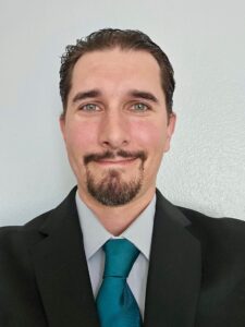 Meet The Staff: A man with short dark hair, a goatee, and light eyes, wearing a black suit jacket, light gray shirt, and teal tie, smiles in front of a plain light-colored wall at Resilience BH.