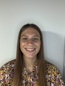 Meet The Staff: A woman with long straight brown hair, wearing a colorful floral shirt, is smiling at the camera against a plain light-colored background—resilience shines through at BH.