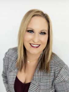 Meet The Staff Resilience BH: A woman with straight blonde hair and blue eyes smiles at the camera. She wears a gray plaid blazer over a burgundy top, with makeup, a nose stud, and a gold necklace, against a plain white background.