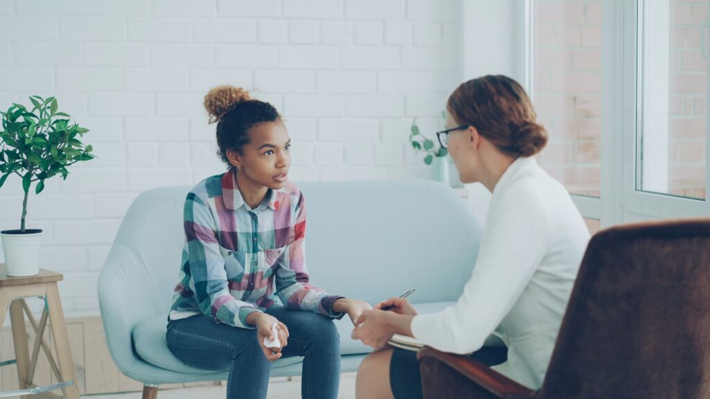 A young girl sits on a sofa, talking and holding tissues, while a woman with a notepad listens attentively—an example of therapies for mental health treatment in a bright, modern office with potted plants and large windows.