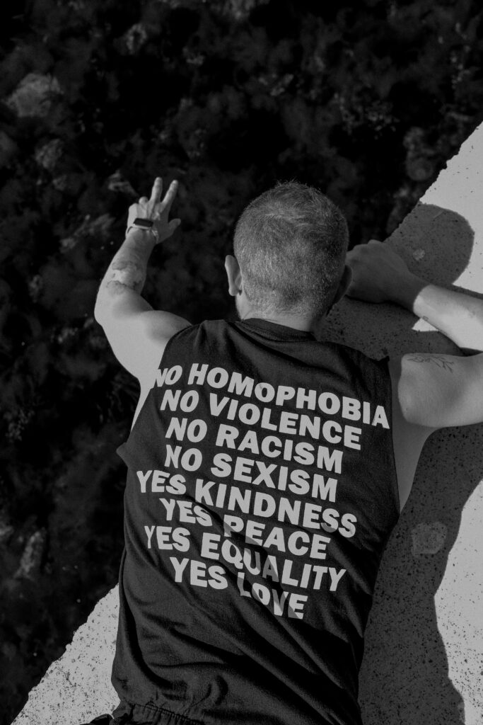 A person lies on a concrete surface, facing away, wearing a shirt with words advocating for equality, kindness, peace, and love—values often promoted in therapies for mental health treatment. The person holds up a peace sign.