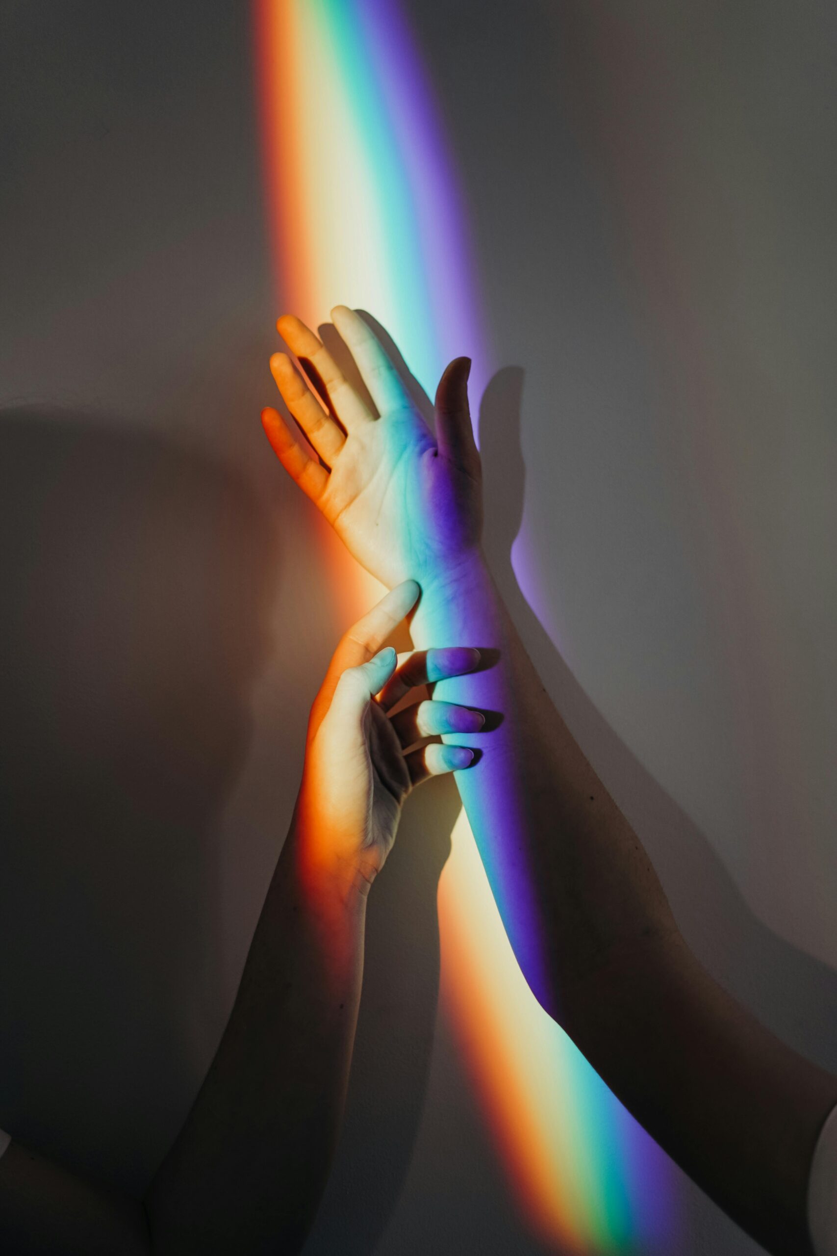 Two hands touch as a rainbow-colored light is cast across them and a plain wall, evoking hope and the healing potential found in therapies for mental health treatment.