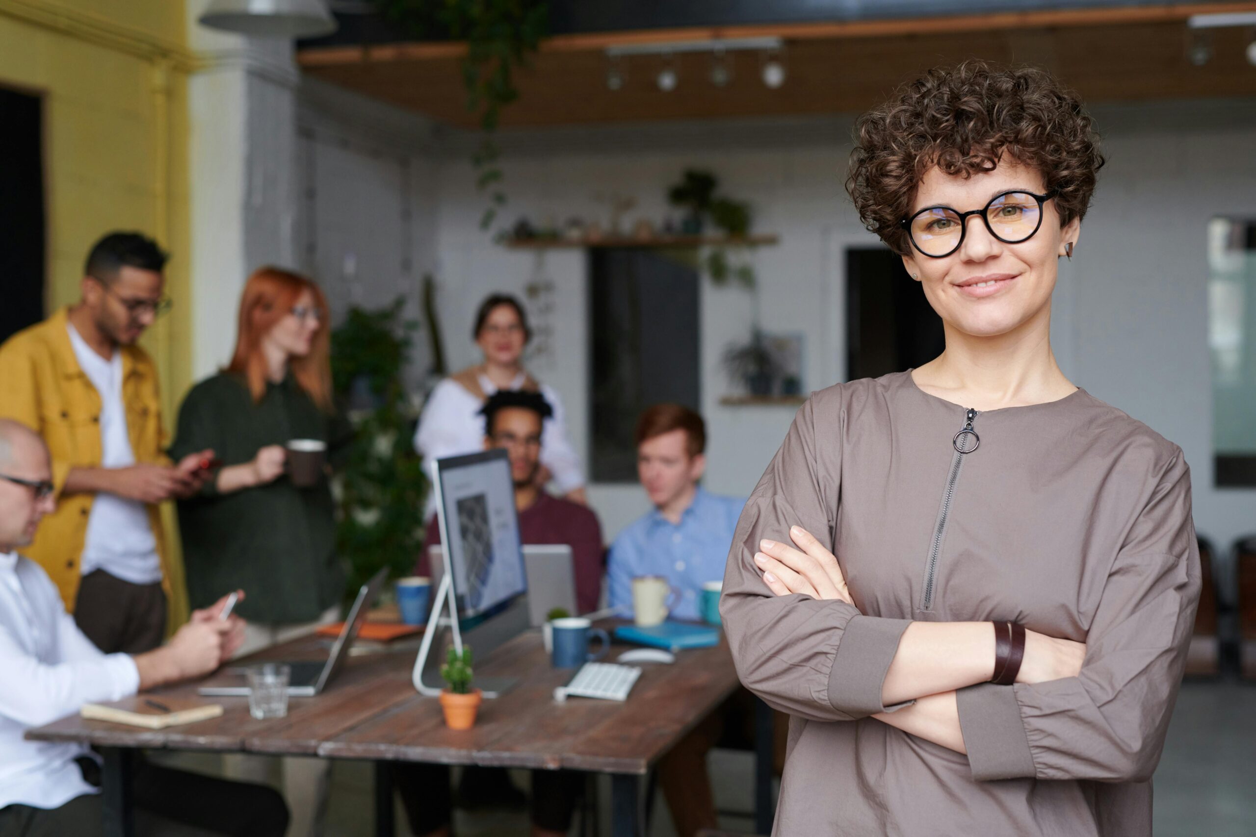A confident person with curly hair and glasses stands smiling with arms crossed in an office, where colleagues discuss therapies for mental health treatment and collaborate around a table in the background.