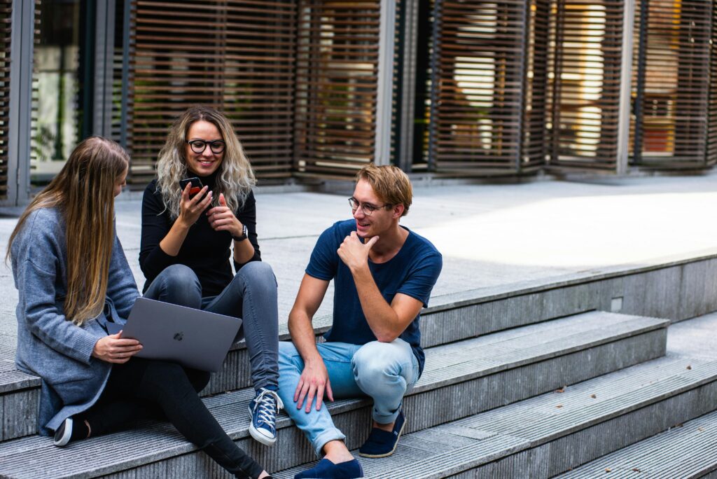 Three young adults sit on outdoor steps, chatting and smiling. As they discuss therapies for mental health treatment, one holds a laptop, another a phone, while the third sits casually—engaged in conversation against a modern building backdrop.
