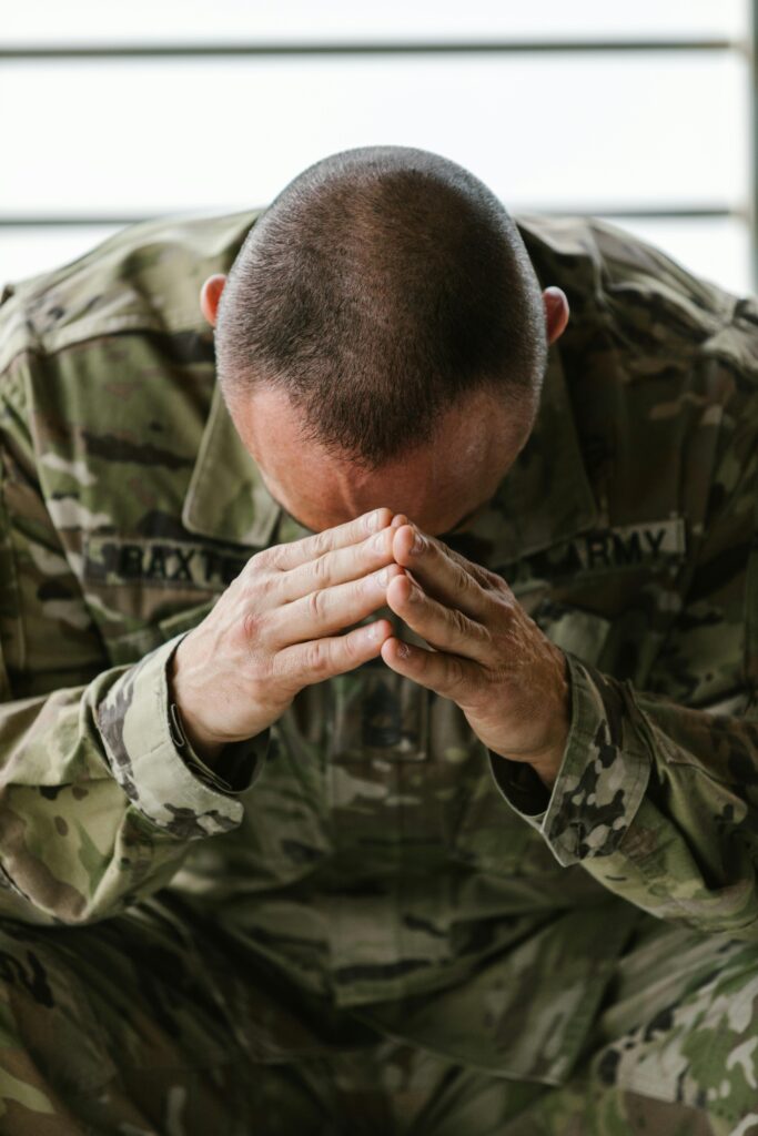 A person in military camouflage uniform sits with elbows on knees, head bowed, and hands pressed together in front of their face, appearing deep in thought or prayer—perhaps reflecting on therapies for mental health treatment.