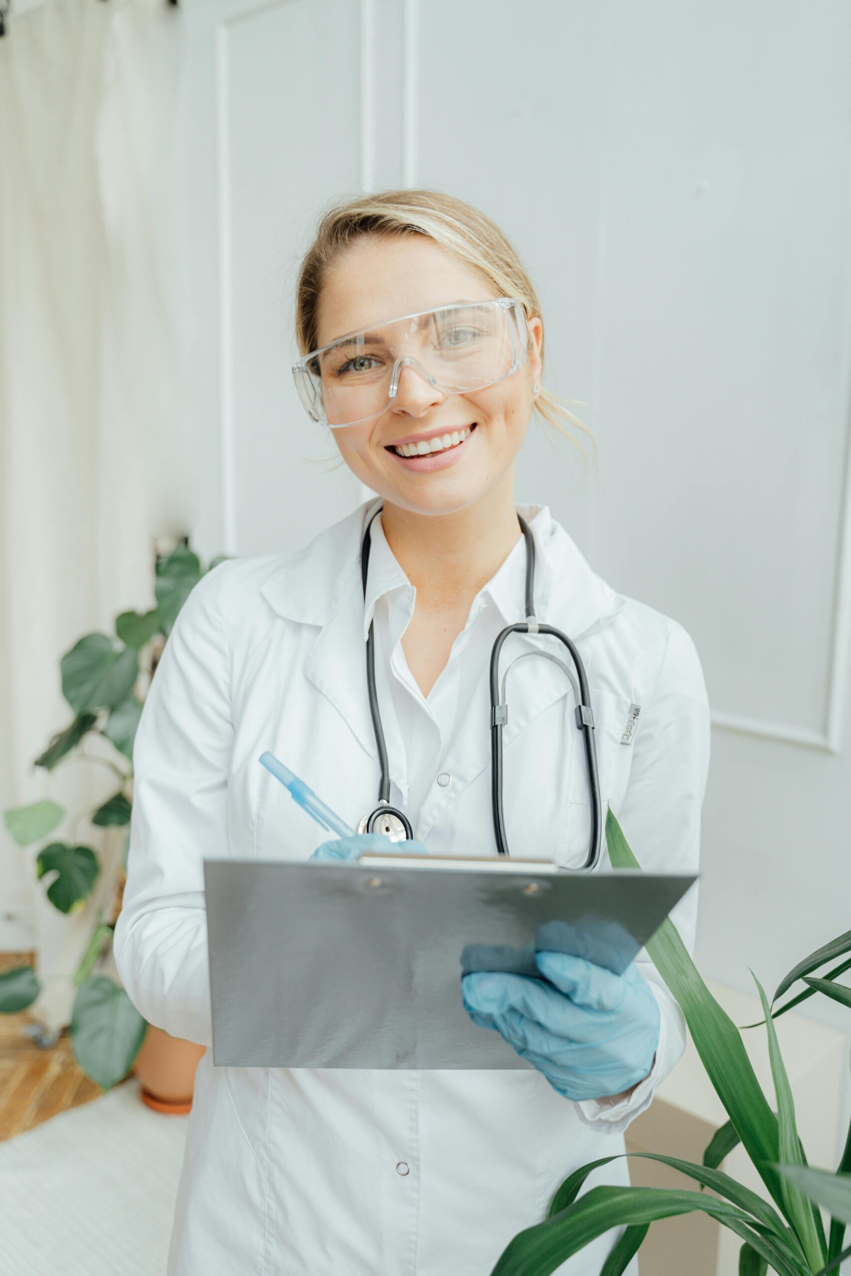 A smiling healthcare professional in a white lab coat, safety glasses, and blue gloves holds a clipboard and pen, standing indoors with plants in the background, ready to discuss therapies for mental health treatment.