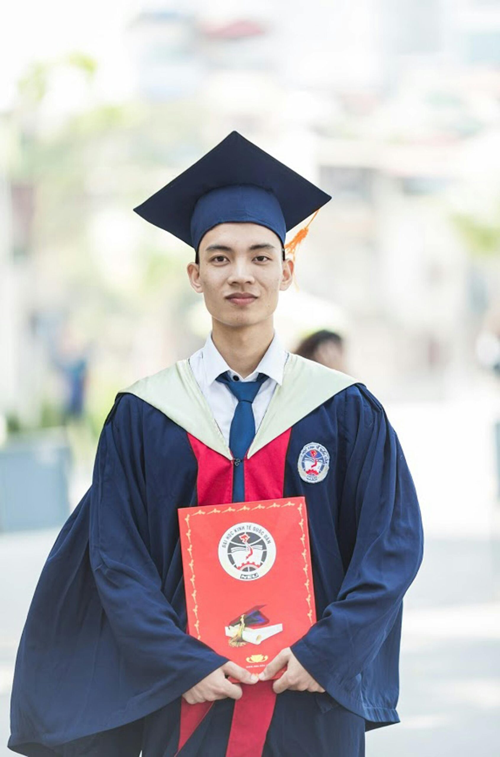 A young man wearing a graduation cap and gown holds a red certificate folder and stands outdoors, looking at the camera with a slight smile—proudly celebrating his studies in therapies for mental health treatment. The background is softly blurred.