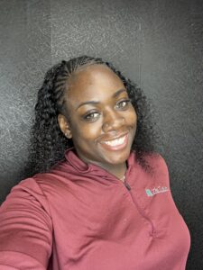 Meet The Staff: A woman with long curly hair smiles at the camera, wearing a maroon zip-up shirt with a small embroidered Resilience BH logo on the chest, standing against a textured dark background.