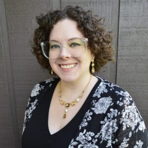 Meet The Staff: A smiling person with short curly hair, wearing clear glasses, a black top, a black floral-patterned cardigan, and matching gold jewelry, stands in front of a textured wooden wall—embodying the resilience of BH.