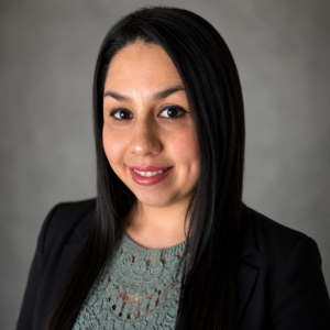 Meet The Staff: A woman with long straight black hair, wearing a black blazer and a green lace top, smiles at the camera against a plain gray background, representing the resilience of the team at Resilience BH.