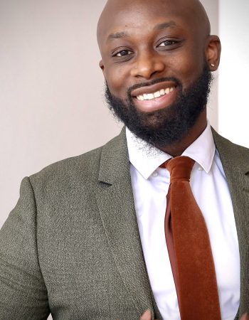 Meet The Staff Resilience BH: A smiling man with a bald head and beard wears a green textured suit, white shirt, and rust-colored tie, adjusting his jacket with one hand. The background is plain and softly lit.